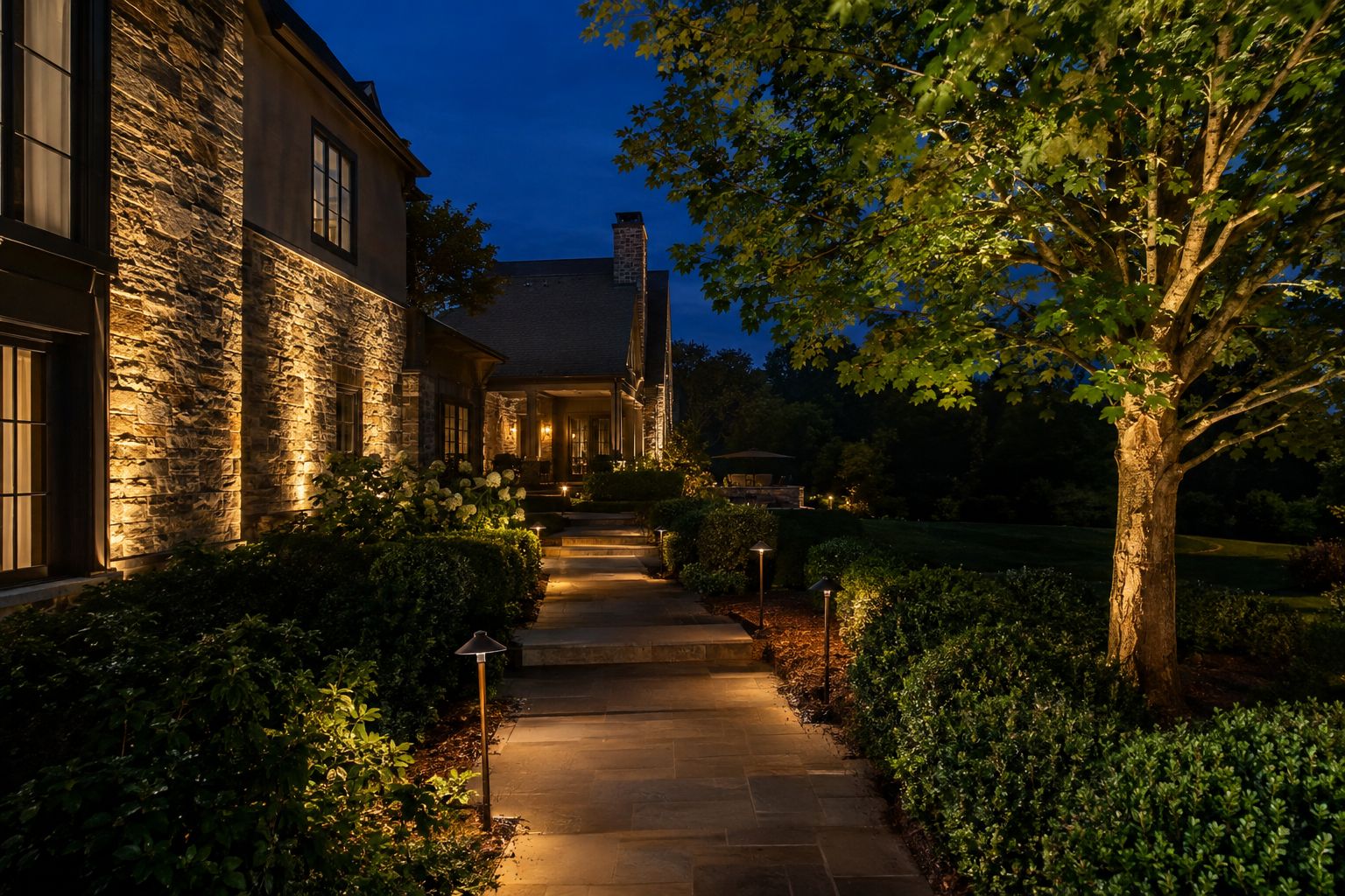 Stone home facade and maple tree lit by warm low-voltage uplights with path lights along a walkway